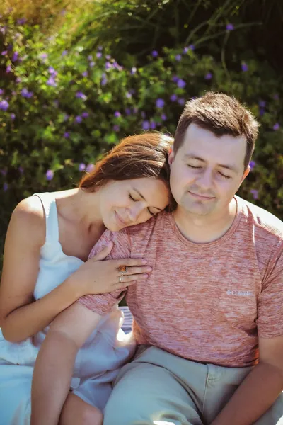 Um casal sentado junto a um arbusto de flores lilás, a cabeça dela encostada ao ombro dele — Olesia Shelkovenko